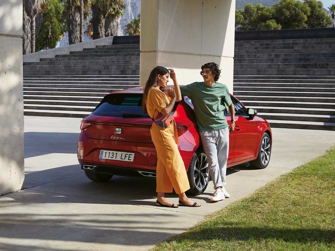 A red SEAT Leon parked under a modern architectural structure. A couple stands nearby, emphasizing the reliability and long-term benefits of SEAT's Service & Maintenance plan for safe and economical vehicle upkeep.