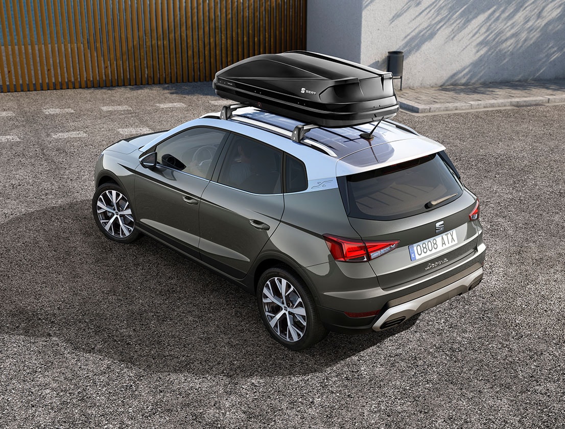 Semi-rear view of the SEAT Arona showcasing a black roof box, to show how the car and the accessory combine. The vehicle is set against a sunny outdoor environment.