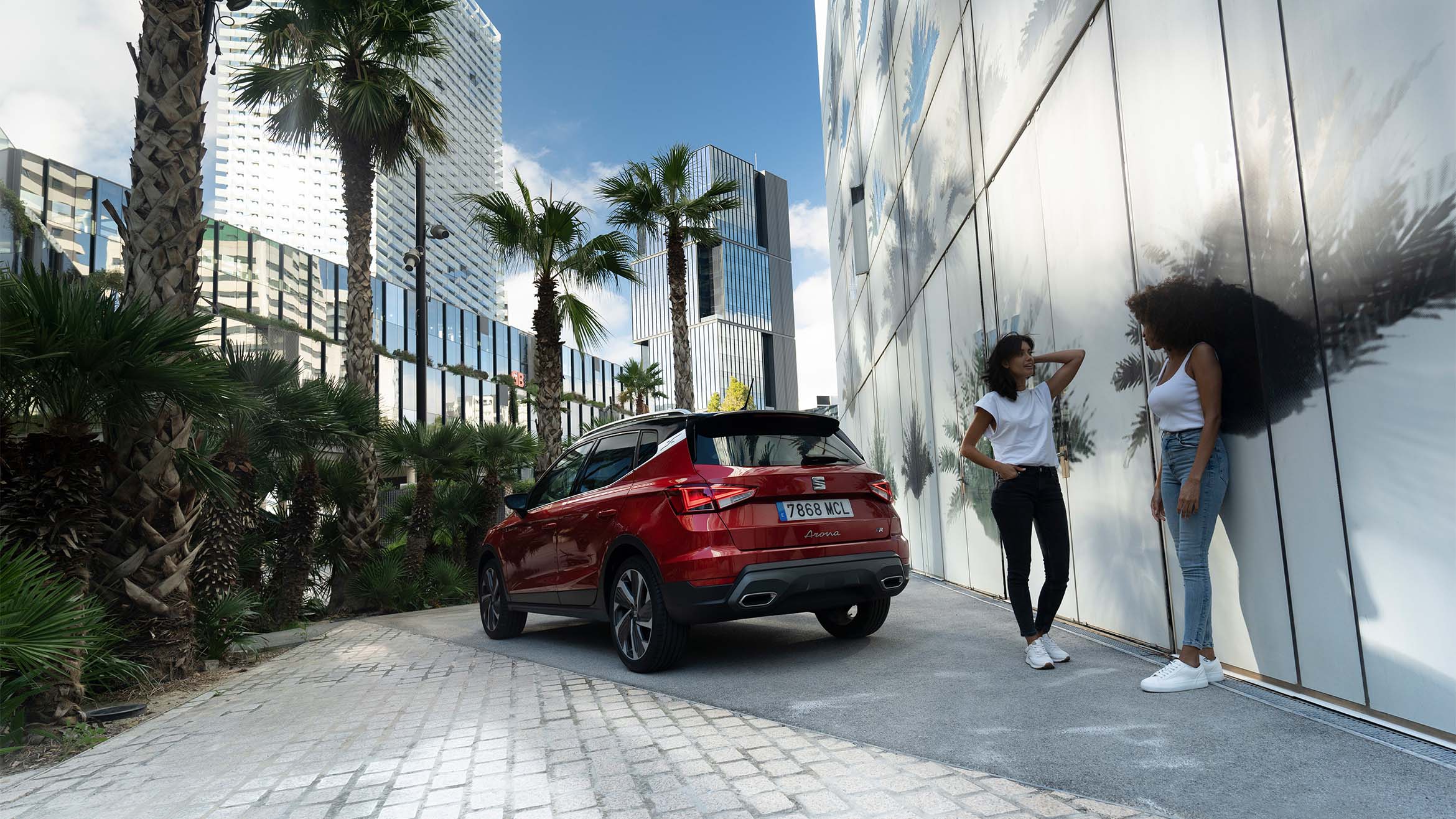 two women talking and laughing to each other in the street next to a SEAT Arona in desire red color