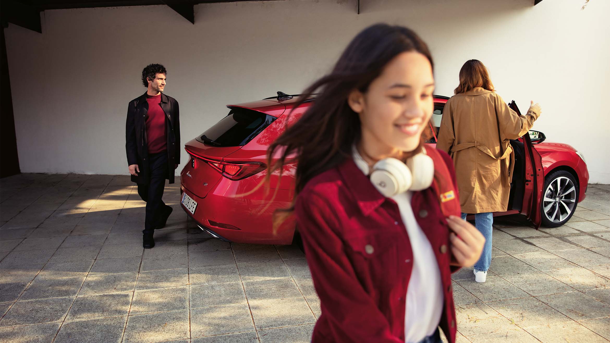 A red SEAT Leon SP Heather OW parked in a driveway with a family around it. The car features sleek, aerodynamic lines, LED taillights and a spacious interior. A man stands near the rear of the car, a woman is opening the rear door and a young girl with headphones is walking in the foreground, highlighting the car's family-friendly design and modern features.
