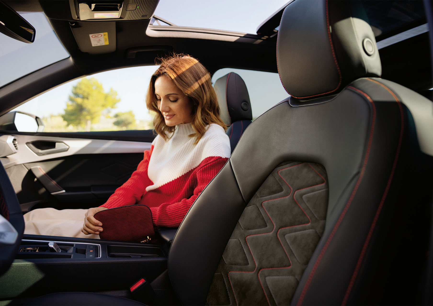 A woman seated inside a SEAT Leon 5D FR, showcasing the car's interior design. The interior features ergonomic seats with red stitching, a sleek dashboard and an advanced infotainment system. The sunroof allows natural light to fill the cabin, highlighting the car's comfortable environment.