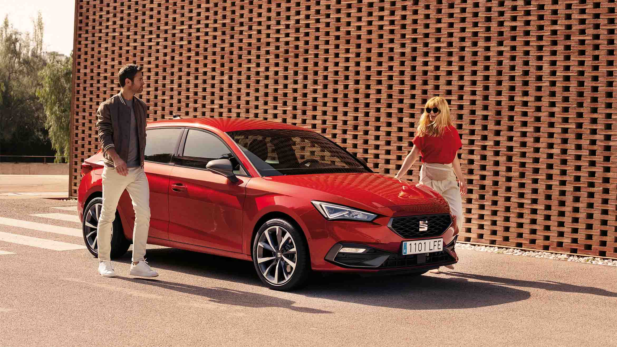 A red SEAT Leon 5D Heather OW parked in front of a brick wall. The car features sleek, aerodynamic lines, LED headlights and alloy wheels. A man and a woman are standing beside the car, both dressed casually. The man wears a brown jacket and white pants, while the woman wears a red top and beige skirt, highlighting the car’s stylish design.