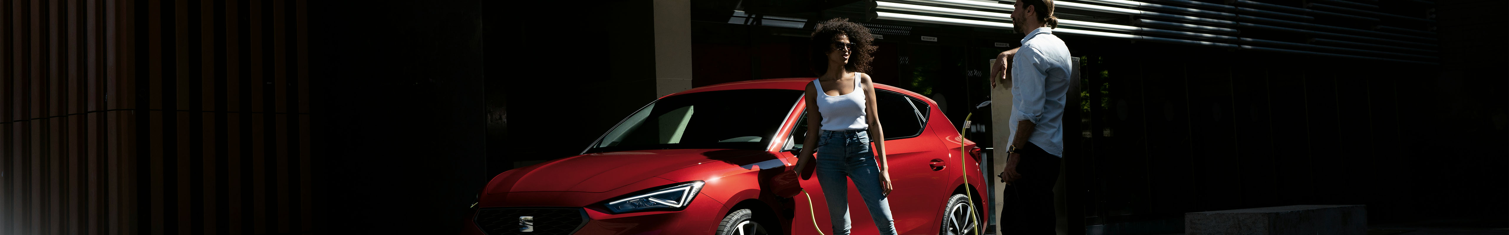 Phev seat leon in red and couple discussing whether its worth buying a hybrid car, low angle and front three quarter view.