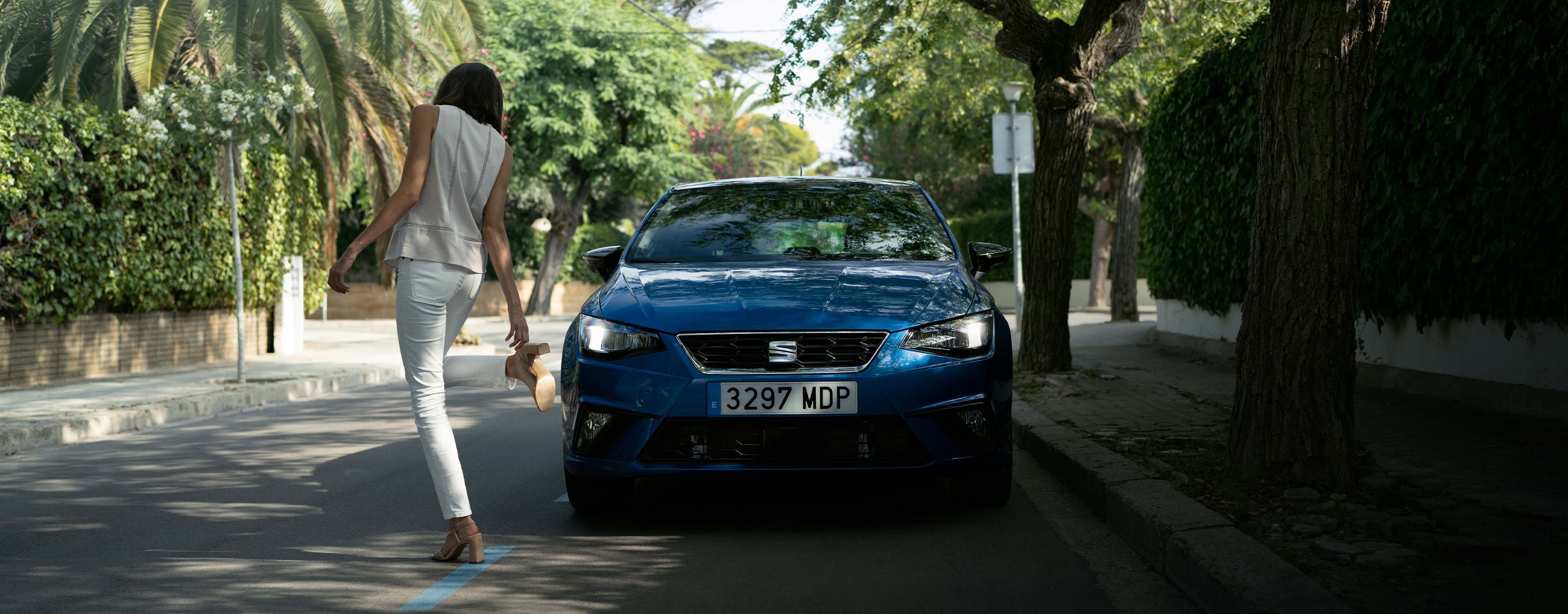 A blue ibiza parked in the street under trees and a woman dressed in white dancing around it.