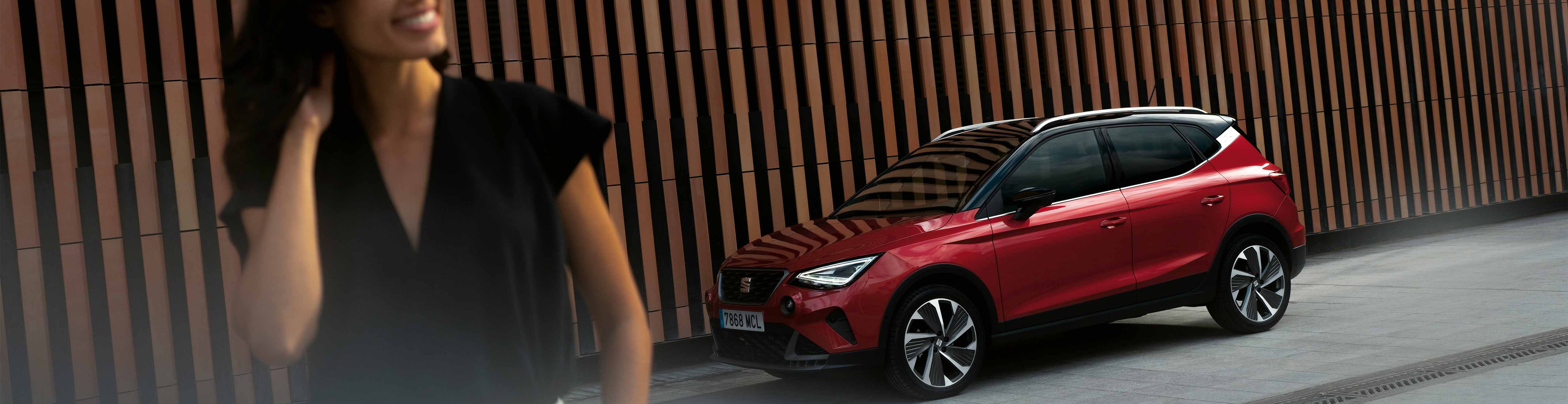 New seat arona in red outside awaiting car servicing and maintenance, woman in foreground smiling.