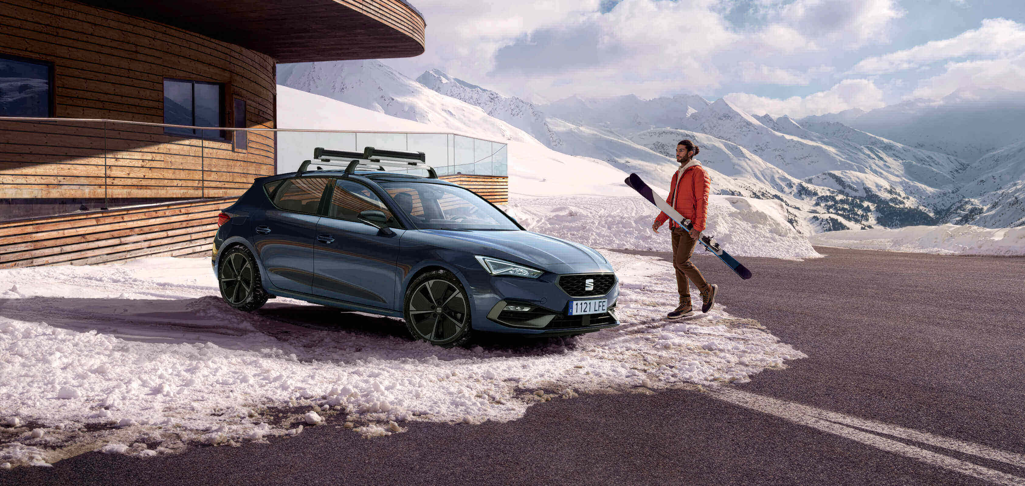 : A grey magnetic tech, side view, parked on snow, by a cabain and a man shoveling snow.