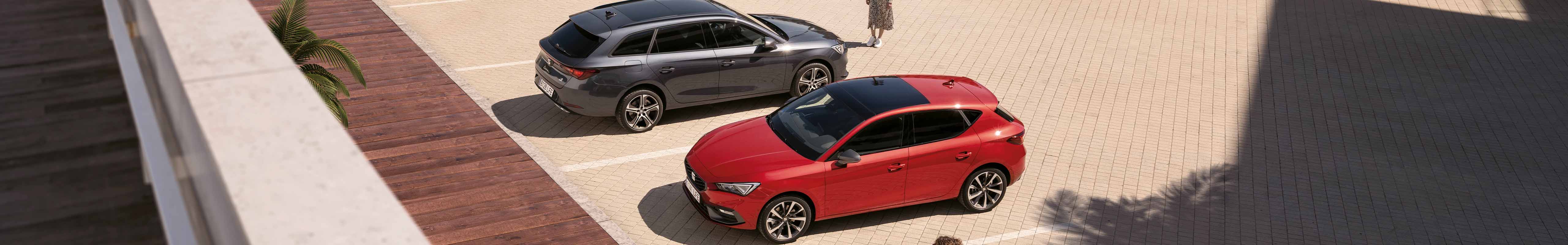  Bird's eye view of two SEAT Leon cars, one in Magnetic Tech Grey and the other in Desire Red, parked with a woman walking by.
