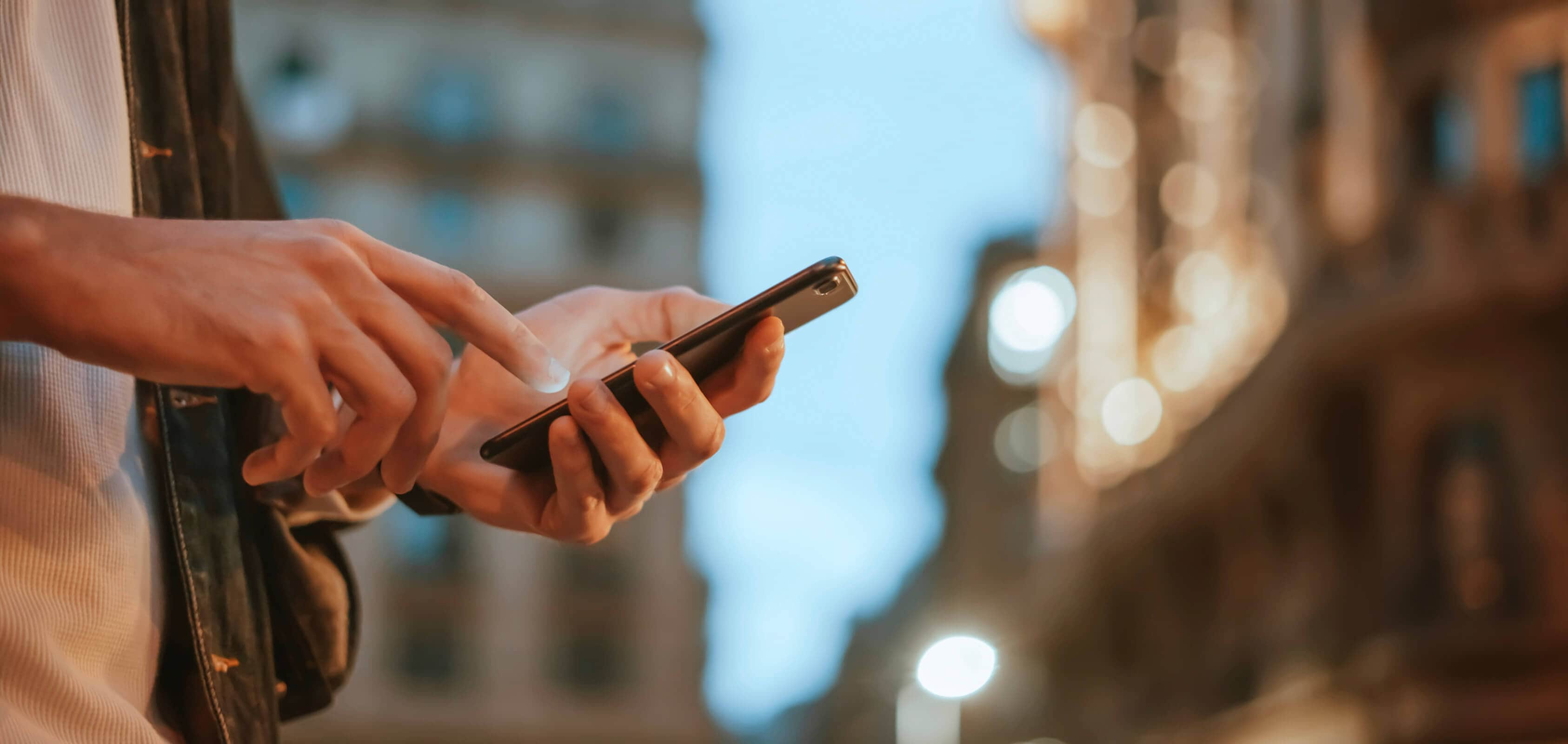 partial view of a man interacting with his phone and the background of the city with buildings and lights. 